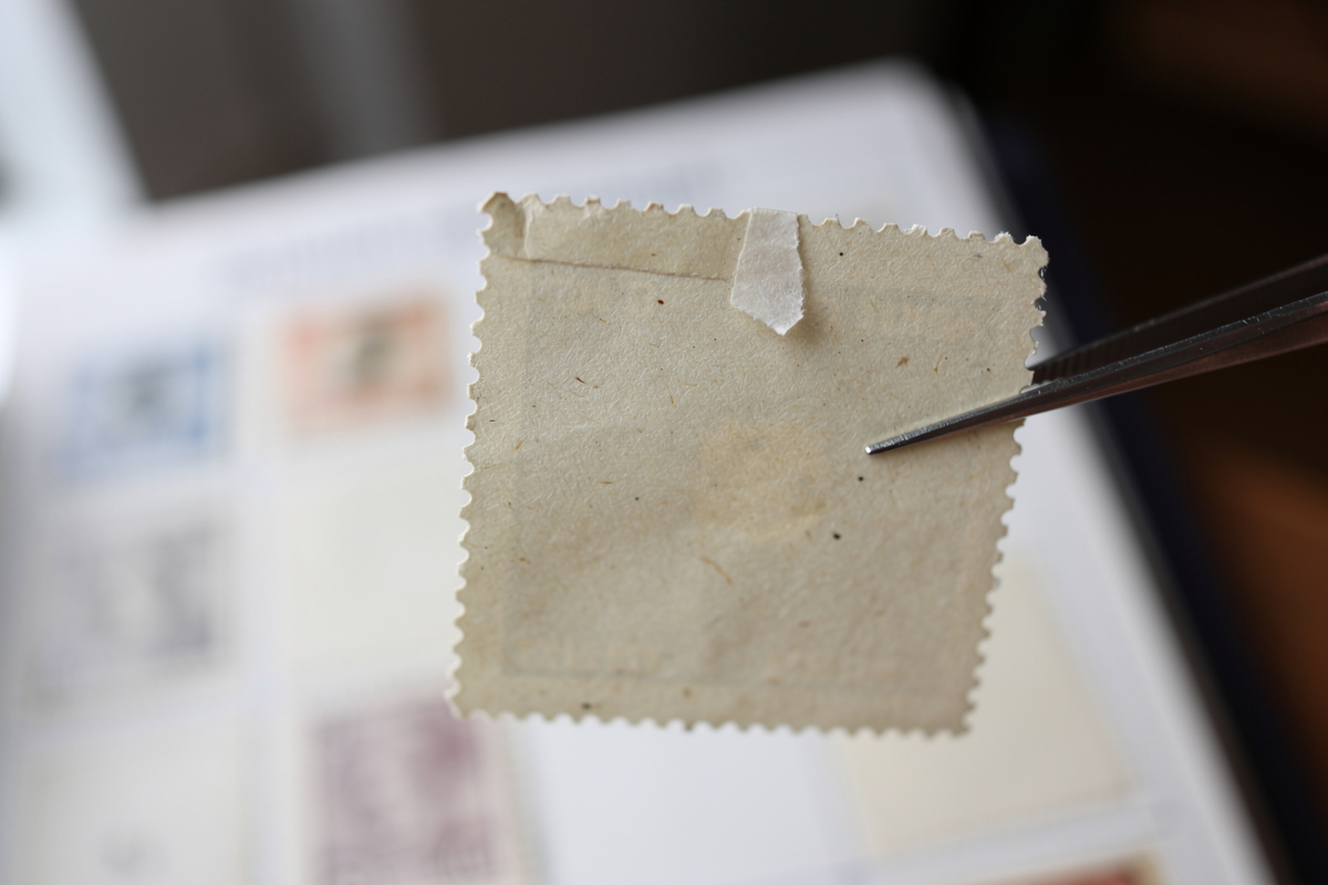 Rückseite einer Briefmarke mit Falzrest in der Pinzette - der Zustand entscheidet über den Marktpreis
